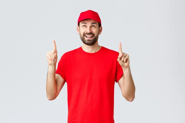 Nätshopping, leverans under karantän- och hämtningskoncept. Glada glada kurir i röd uniform mössa och t-shirt, leende förvånad och peka fingrar upp, läsa banner eller tecken — Stockfoto