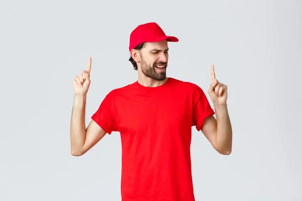 Upbeat stilig skäggig leverans kille spendera paus avkopplande, anställd i röd uniform mössa och t-shirt sång, peka finger upp sorglös, blunda, stående grå bakgrund — Stockfoto