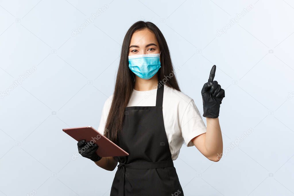 Covid-19, online orders, small coffee shop and preventing virus concept. Smiling friendly asian waitress, female barista in medical mask and gloves point upper right corner, use digital tablet