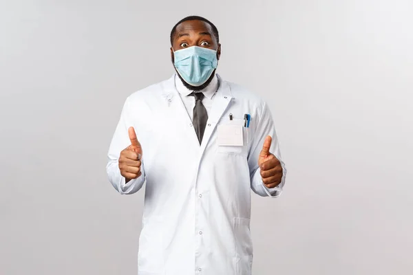 Portrait de joyeux, excité afro-américain médecin masculin en manteau blanc, recommander une médecine impressionnante ou un service clinique, montrer pouces levés avec sourire heureux étonné, garantir la meilleure qualité — Photo