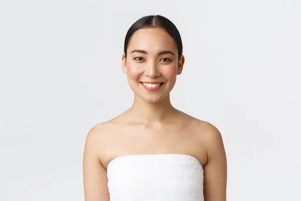 Affascinante sorridente giovane donna asiatica in asciugamano guardando la fotocamera felice come finire la sua routine quotidiana di cura della pelle, in piedi bagno su sfondo bianco, andando a doccia, frequentare salone spa — Foto Stock