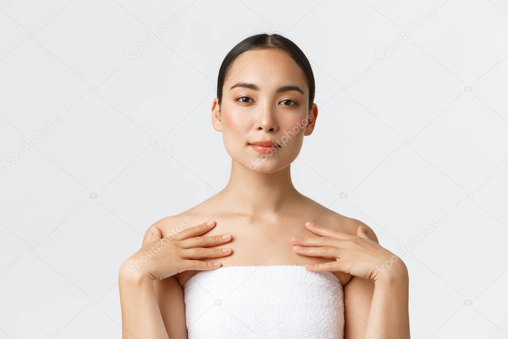 Tender beautiful asian woman with clean skin, perfect face, standing in towel and touching body, applying cream or cleanser, skincare products in bathroom, attend spa salon, white background