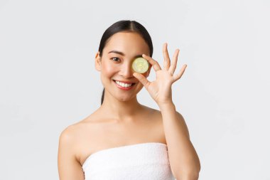 Güzellik, kişisel bakım, kaplıca salonu ve cilt bakımı konsepti. Banyo havlusu içinde güzel bir Asyalı kadın salatalık parçasını gözünün üstünde tutuyor ve gülümsüyor. Nemlendirici yüz maskesi ya da temizleyici, duş jeli...