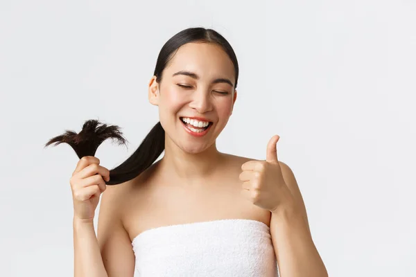 Beauty, prodotti per la perdita dei capelli, shampoo e concetto di cura dei capelli. Primo piano di soddisfatto, felice ragazza asiatica in asciugamano da bagno che mostra pollici-up e capelli sani estremità, in piedi soddisfatto sfondo bianco — Foto Stock