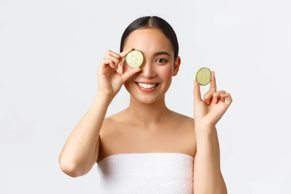 Szépség, testápolás, spa szalon és bőrápolás koncepció. Gyönyörű fiatal ázsiai nő fürdőtörölköző gazdaság uborka és mosolygós, promo arc- vagy testkezelés, hidratáló jellemzői krém — Stock Fotó