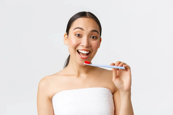 Piękno, osobista opieka i higiena. Entuzjastyczna szczęśliwa azjatycka kobieta w ręczniku kąpielowym przygotowuje się, myje białe zęby, trzyma szczoteczkę do zębów i uśmiecha się szeroko, białe tło — Zdjęcie stockowe