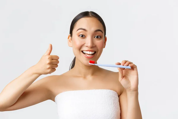 Kecantikan, perawatan pribadi, dan konsep kebersihan. Wanita asia yang menarik dalam handuk mandi mencoba sikat gigi baru, menunjukkan jempol pasta gigi besar untuk pemutihan gigi, latar belakang putih — Stok Foto