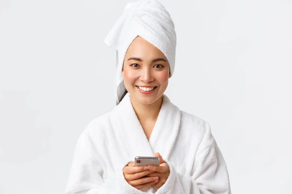 Cura personale, bellezza delle donne, bagno e doccia concetto. Primo piano di bella giovane donna asiatica si prepara, in piedi in asciugamano da bagno e accappatoio con il telefono cellulare, fotocamera sorridente — Foto Stock