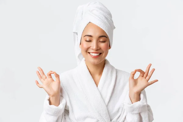 Cura personale, bellezza delle donne, bagno e doccia concetto. Primo piano di sollevato attraente donna asiatica partecipare spa salone di bellezza, indossare asciugamano da bagno e accappatoio, meditando, sensazione di nirvana, sorridente — Foto Stock