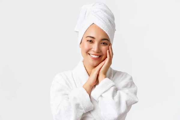 Cuidados pessoais, mulheres beleza, banho e chuveiro conceito. Close-up de bela mulher asiática feliz em toalha e roupão tocando rosto suavemente, sorrindo dentes brancos, promo de cuidados com a pele e produtos de higiene — Fotografia de Stock