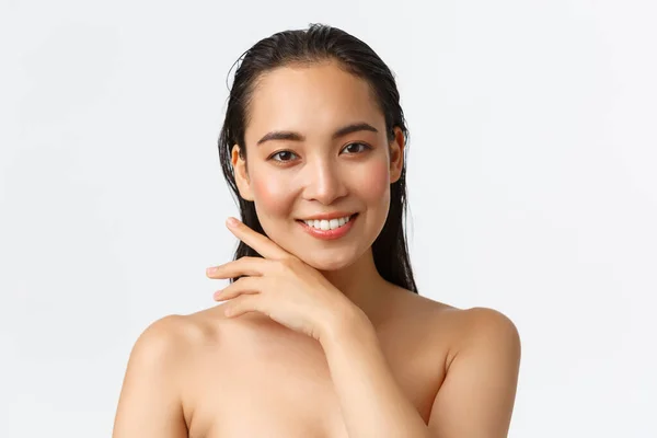 Hudvård, kroppsvård, skönhet och badkoncept. Närbild av sensuell attraktiv asiatisk kvinna som står naken i dusch, ler och försiktigt röra ansiktet, främjande av ansiktsprodukter, vit bakgrund — Stockfoto