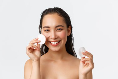 Cilt bakımı, kadın güzelliği, hijyen ve kişisel bakım konsepti. Güzel Asyalı bir kızın çıplak durduğu ve yüzünün yanında pamuk pedleri olan duştan önce makyajını silen, beyaz arka plan.