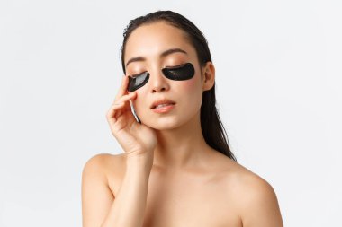 Skincare, women beauty, hygiene and personal care concept. Close-up of sensual pretty asian female standing naked in bathroom, apply under eye patches, eye mask for dark circles and puffiness clipart