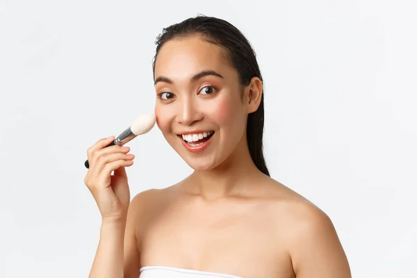 Skincare, kecantikan wanita, kebersihan dan konsep perawatan pribadi. Close-up of excited asian female applying makeup with brush, looking at camera and smile gused, white background — Stok Foto