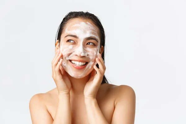 Cura della pelle, bellezza femminile, igiene e cura personale. Primo piano di bella donna asiatica nuda sorridente e guardando a sinistra ottimista mentre la pelle pulita con schiuma detergente, prodotto per la cura quotidiana — Foto Stock