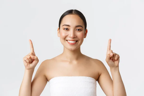 Kecantikan, kosmetologi dan konsep salon spa. Wanita asia yang gembira dengan handuk tersenyum puas dan menunjuk jari ke atas, merekomendasikan terapi pijat, produk perawatan kulit, latar belakang putih — Stok Foto