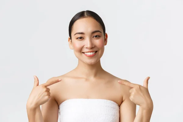 Beauty, cosmetology and spa salon concept. Close-up of satisfied beautiful asian female client in white towel smiling and pointing at herself, recommend beauty clinic and skincare procedures — Stock Photo, Image