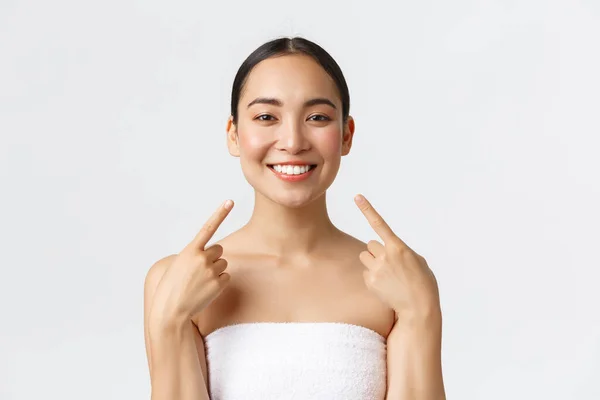 Beauty, cosmetology and spa salon concept. Close-up of happy asian woman with white perfect teeth, clean skin pointing at face with pleased grin, satisfied with cosmetic procedures in beauty clinic — Stock Photo, Image