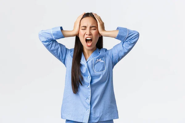 Distressed pissed-off asian girl in blue pajamas screaming outraged, holding hands on head in denial, got huge trouble, feeling upset and disappointed, standing white background — Stock Fotó