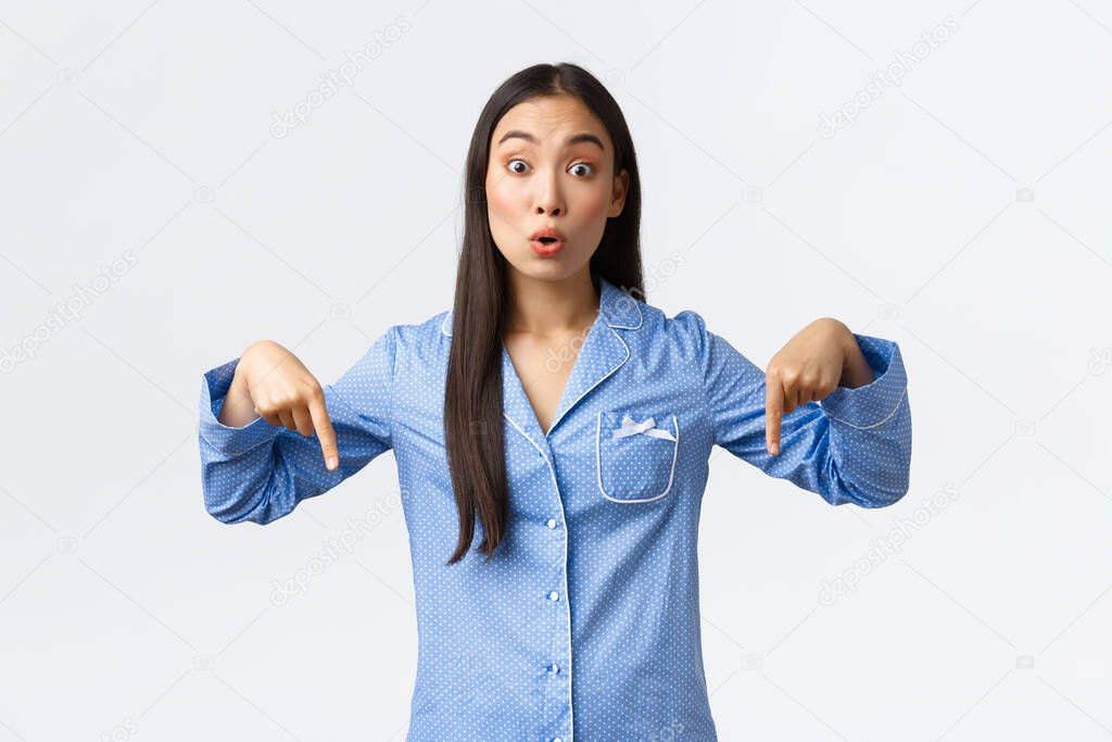 Surprised and curious beautiful korean girl in blue pajamas asking question and looking intrigued at camera as pointing fingers down. Female girlfriend at sleepover party found something interesting