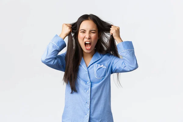 Indignada y cabreada chica coreana en pijama azul tirando el pelo de la cabeza y gritando enojado, de pie frustrado, sintiéndose harto y deprimido, de pie molesto sobre el fondo blanco — Foto de Stock