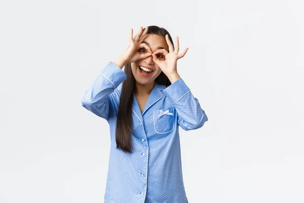 Feliz chica asiática encantadora en pijamas azules mostrando gestos de gafas falsas y sonriendo optimista, divirtiéndose con los hermanos en la fiesta de pijamas, usando pijamas y tontos, fondo blanco — Foto de Stock