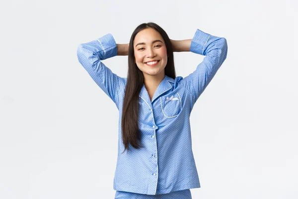 Gülümseyen, mutlu ve rahat Asyalı kız mavi pijamalarıyla mutlu görünüyor, kafasının arkasında uzanmış, tatmin olmuş, rahatlamış ve mutlu bir şekilde beyaz arka planda yatıyor. — Stok fotoğraf