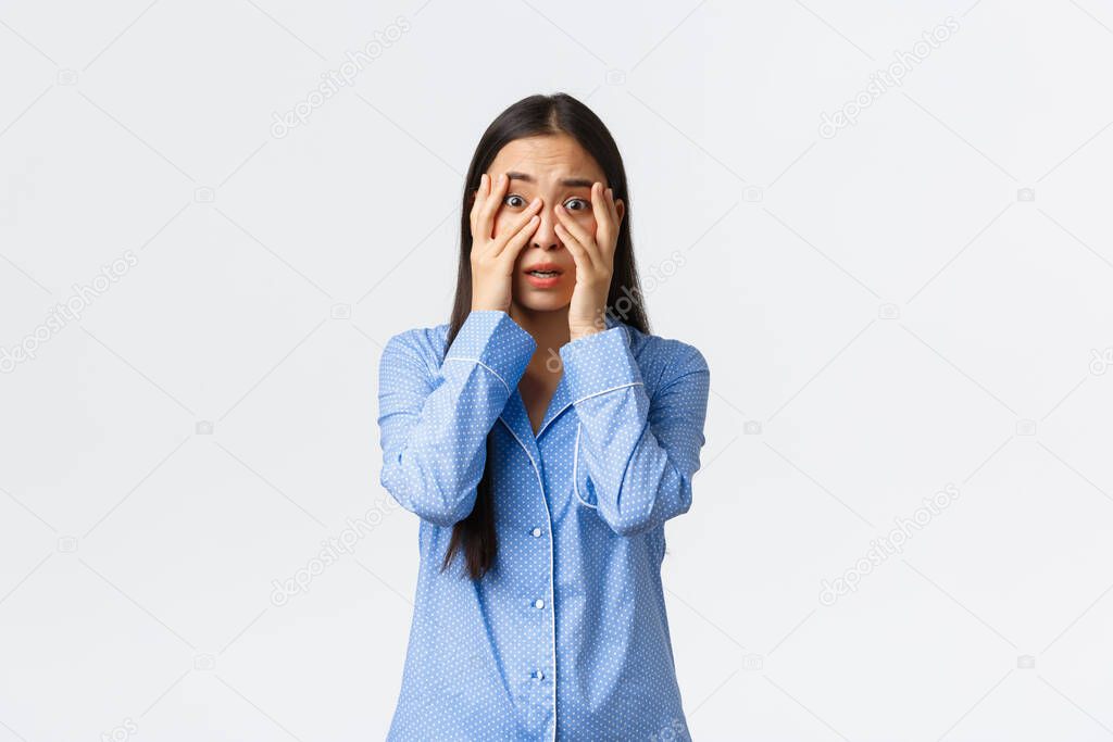 Scared insecure and timid asian girl in blue pajamas shivering, looking through fingers at something scary, watching horror movie during sleepover, standing white background