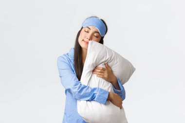 Göz maskesi ve pijamasıyla uyuyan çekici Asyalı kız, yastığa sarılıyor ve gözleri kapalı uzanıyor, rüya görüyor, gece uykusu çekiyor, beyaz arka planda rüya gibi poz veriyor.