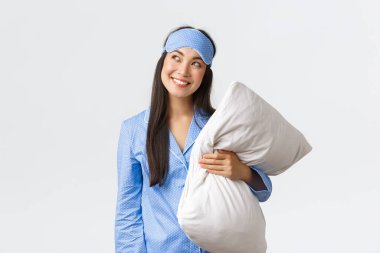 Mavi pijama ve uyku maskesi takmış kurnaz ve düşünceli kawaii Asyalı kız, elinde yastık, sol üst köşede meraklı bakıyor, bir fikri varmış gibi sırıtıyor, beyaz bir arka plan hayal ediyor.