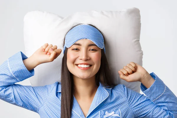 Zbliżenie szczęśliwej i zadowolonej uśmiechniętej azjatki w niebieskiej piżamie i śpiącej masce leżącej w łóżku na poduszce, rozciągającej się zadowolonej i uśmiechniętej po dobrym śnie, budzącej się naładowanej energią — Zdjęcie stockowe