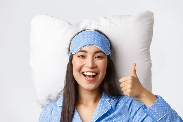 Zbliżenie zadowolonej Azjatki w niebieskiej piżamie i masce sypialnej, leżącej w łóżku na miękkiej wygodnej poduszce, z kciukami do góry w aprobacie, polecam produkt lub tabletki bezsenne — Zdjęcie stockowe