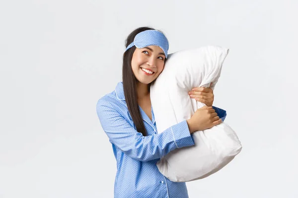 Dreamy beautiful asian girl in blue pajamas and sleeping mask hugging pillow and smiling pleased as looking upper left corner imaging something in bed, picturing, using imagination — Stock Photo, Image