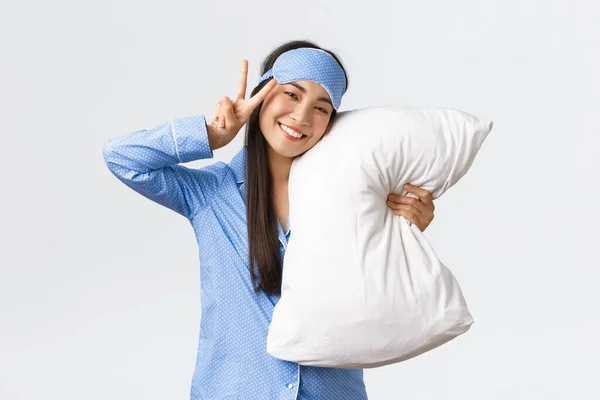 Kawaii glad leende asiatisk flicka i blå sovmask och pyjamas, krama kudde och visar fred tecken, mår bra efter god natts sömn, ligger i sängen under övernattning, vit bakgrund — Stockfoto