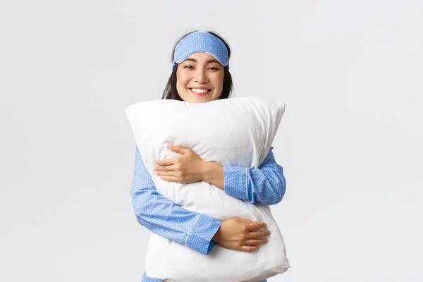 Uśmiechnięta Azjatka w masce do spania i piżamie przytulająca miękką i wygodną poduszkę z usatysfakcjonowanym relaksującym wyrazem twarzy, kładąca się do łóżka, czująca się przytulnie na białym tle — Zdjęcie stockowe