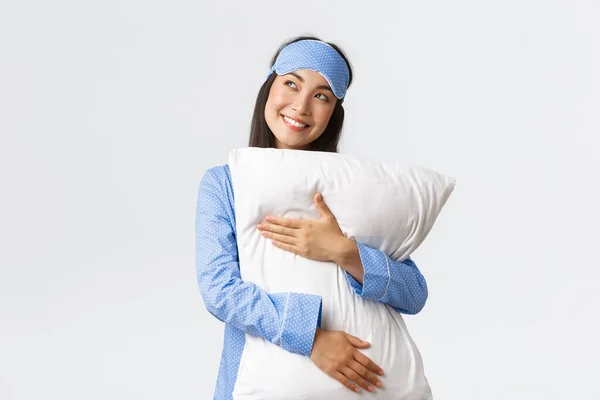 Dromerig mooi aziatisch meisje in blauwe pyjama en slaapmasker knuffelen kussen en glimlachen blij als kijken linksboven beeldvorming iets in bed, beeld, met behulp van verbeelding — Stockfoto