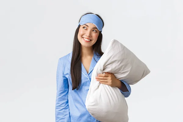 Cunning and thoughtful kawaii asian girl in blue pajama and sleeping mask, holding pillow and looking curious upper left corner, smiling sly as having idea, imaging something, white background — Stock Photo, Image