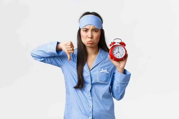 Decepcionado pouting e incomodado bonito ásia menina no azul pijama e dormir máscara queixando-se, ódio acordar cedo para alarme relógio, mostrando ele e polegares para baixo, sulking descontente, branco fundo — Fotografia de Stock