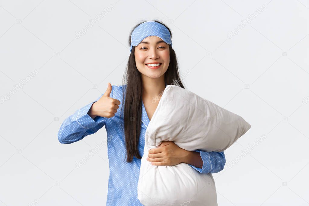 Cheerful pleased cute asian girl in blue pajama and sleeping mask, holding soft comfortable pillow and showing thumbs-up satisfied, had good night sleep, taking pills from insomnia