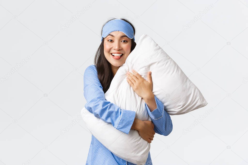 Funny and cute asian girl with happy smile, showing tongue as hugging pillow at sleepover party, wearing pajamas and sleeping mask, fooling around at bed over white background