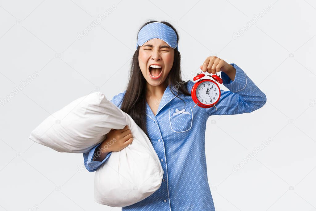Annoyed and mad asian girl in blue pajamas and sleeping mask screaming frustrated as overslept, showing alarm clock and shouting bothered, being late for work, holding pillow