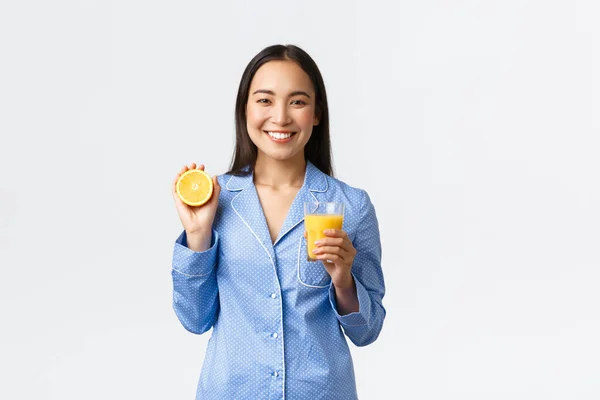 Morning, active and healthy lifestyle and home concept. Smiling cheerful asian girl starting her day with fresh made orange guice, holding glass and half of orange, looking happy and energized