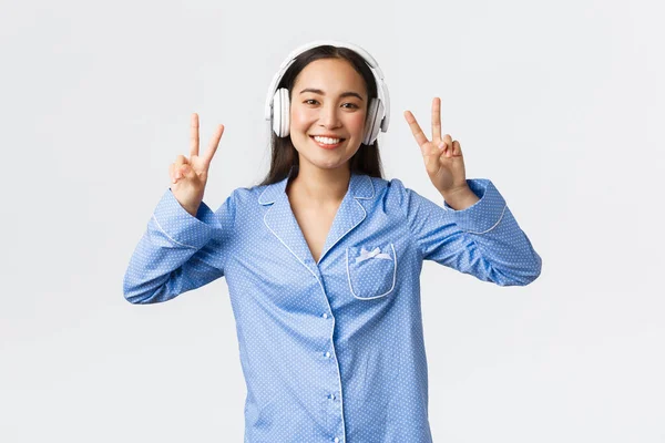Loisirs à domicile, week-ends et concept de style de vie. Kawaii asiatique fille en pyjama écouter de la musique dans les écouteurs sans fil et montrant geste de paix avec un large sourire heureux, fond blanc — Photo