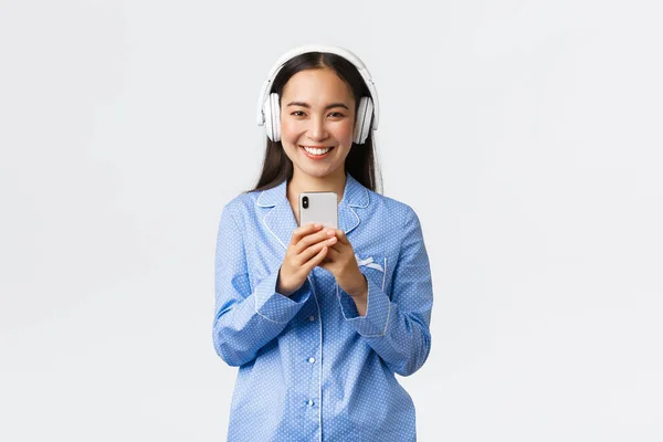 Loisirs à domicile, week-ends et concept de style de vie. Souriant jolie fille asiatique en pyjama bleu écouter de la musique écouteurs sans fil et de prendre des photos sur téléphone mobile, debout fond blanc — Photo