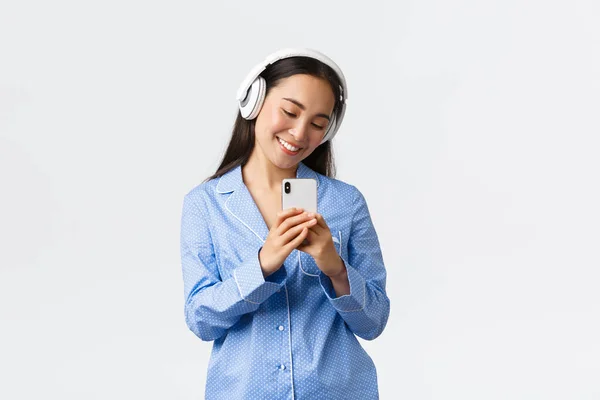 Otthoni szabadidő, hétvégék és életmód koncepció. Gyönyörű női blogger fülhallgatóban és pizsamában, amint szelfit készít a tükörben, és valami aranyosat fényképez a mobiltelefonján. — Stock Fotó