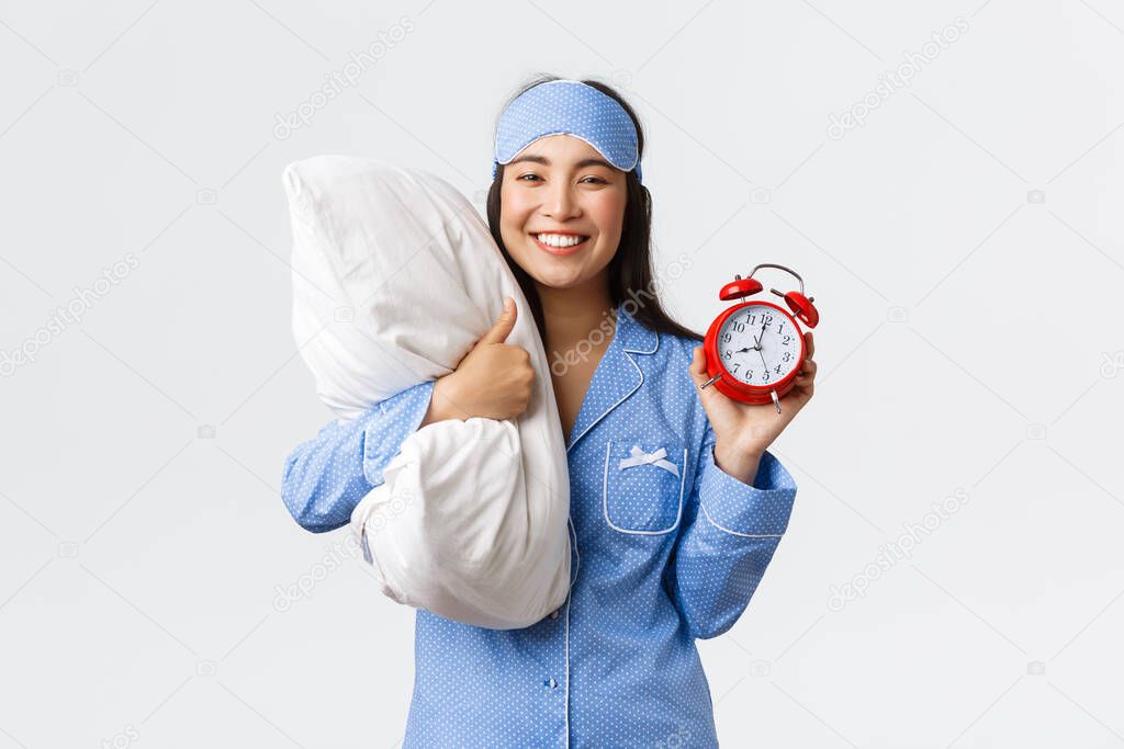 Enthusiastic and happy smiling asian girl in blue pajamas and sleeping mask, showing alarm clock and thumbs-up in approval, like waking up early for morning run, holding pillow