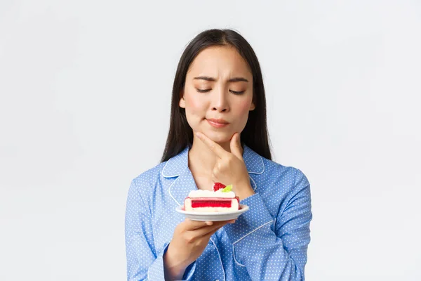 Close-up tiro de ponderando sério-olhando menina asiática no pijama olhando perplexo no bolo como pensar o quanto de calorias na sobremesa, querendo comê-lo à noite, de pé fundo branco — Fotografia de Stock