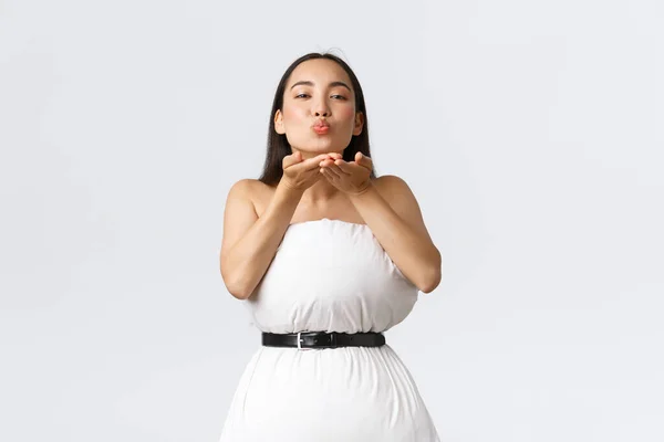 Beauty, Mode und Social Media Konzept. Wunderschöne asiatische Frau macht Kleid aus Kissen mit Gürtel um Abfall gesichert, pustet Luftkuss vor der Kamera kokett über weißem Hintergrund — Stockfoto