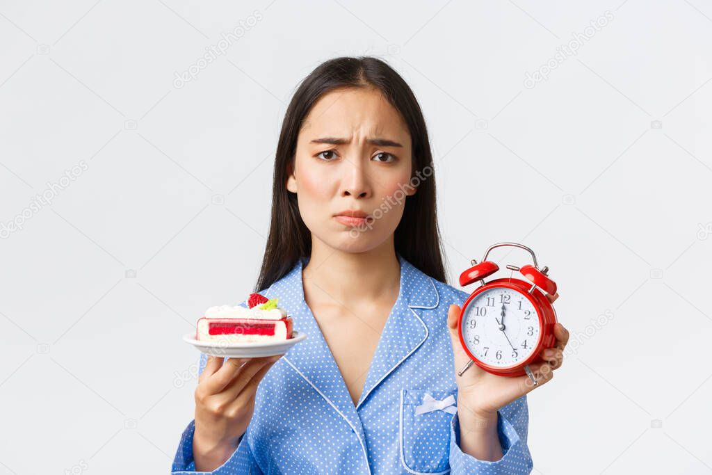 Close-up shot of upset silly asian girl on diet, showing clock and tasty piece of cake, pouting as want to eat dessert but its too late night, standing white background displeased