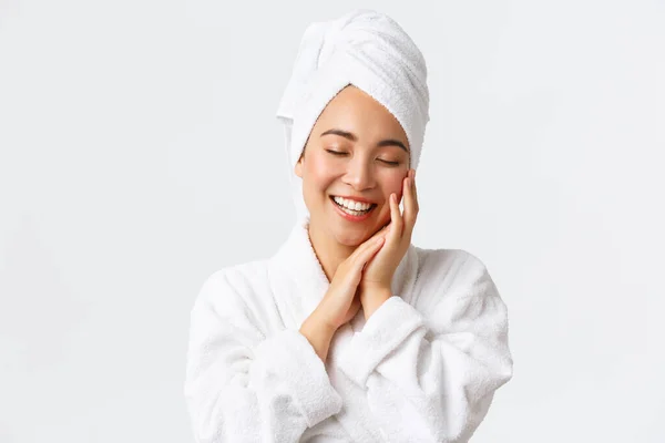 Cuidados pessoais, mulheres beleza, banho e chuveiro conceito. Close-up de bela mulher asiática feliz em toalha e roupão tocando rosto suavemente, sorrindo dentes brancos, promo de cuidados com a pele e produtos de higiene — Fotografia de Stock